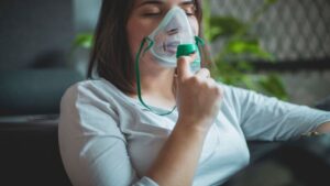A Young Brunette Is Sitting on the Couch and Using an Inhaler to Make Her Breathing Easier